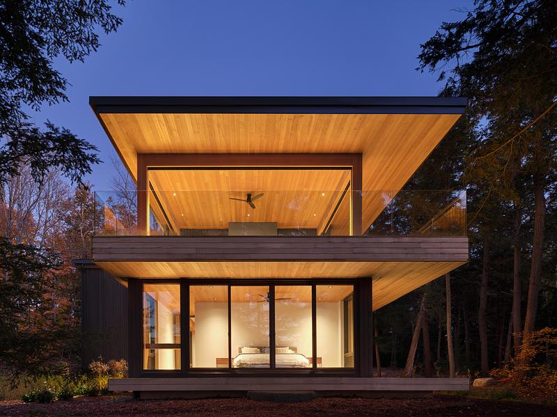 Maison contemporaine en bois avec grandes baies vitrées éclairée de nuit en pleine forêt au Canada