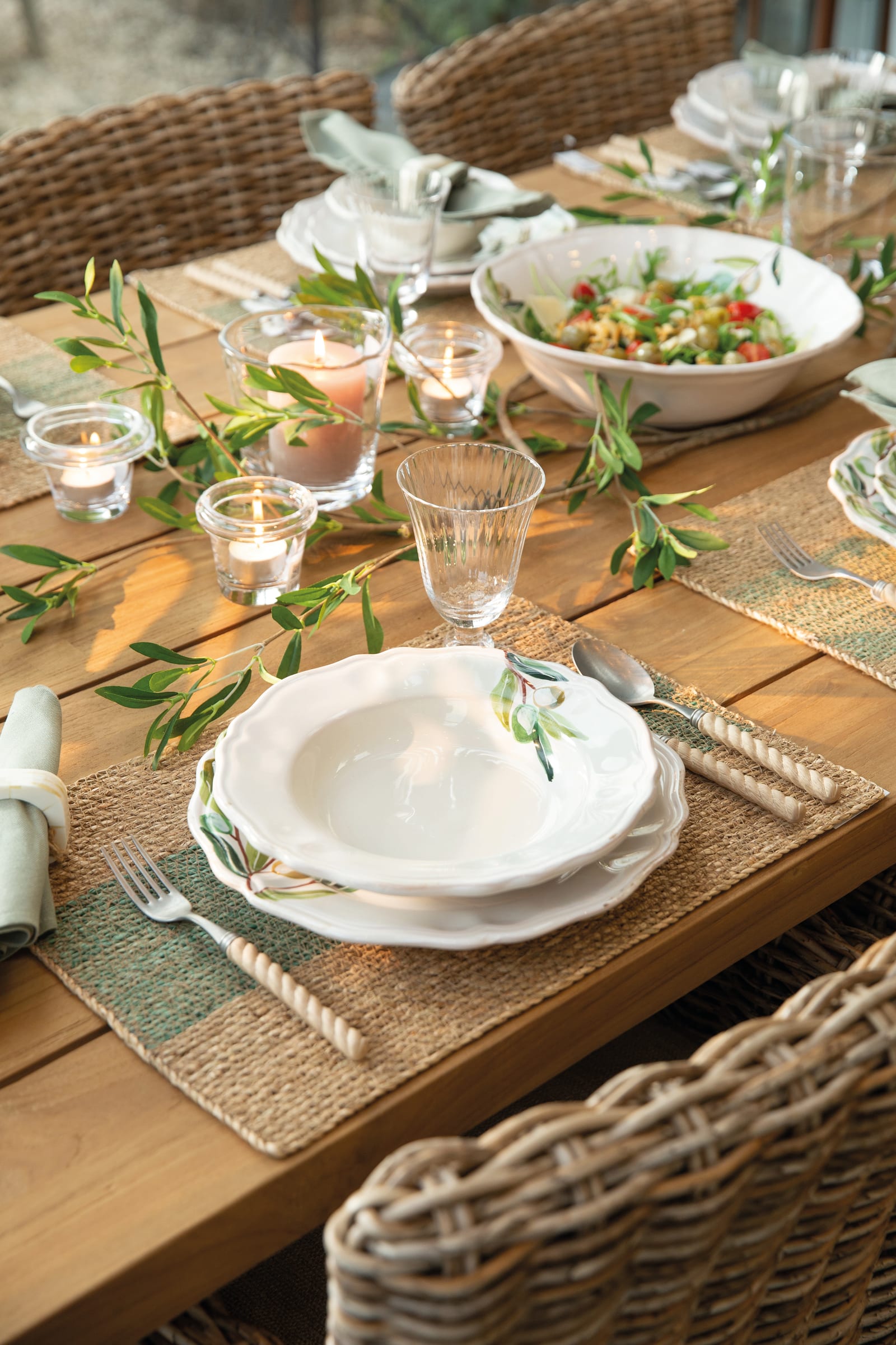Assiettes en céramique blanche décorées d'olives sur un set de table en jonc, bougies chauffe-plat et branches d'olivier