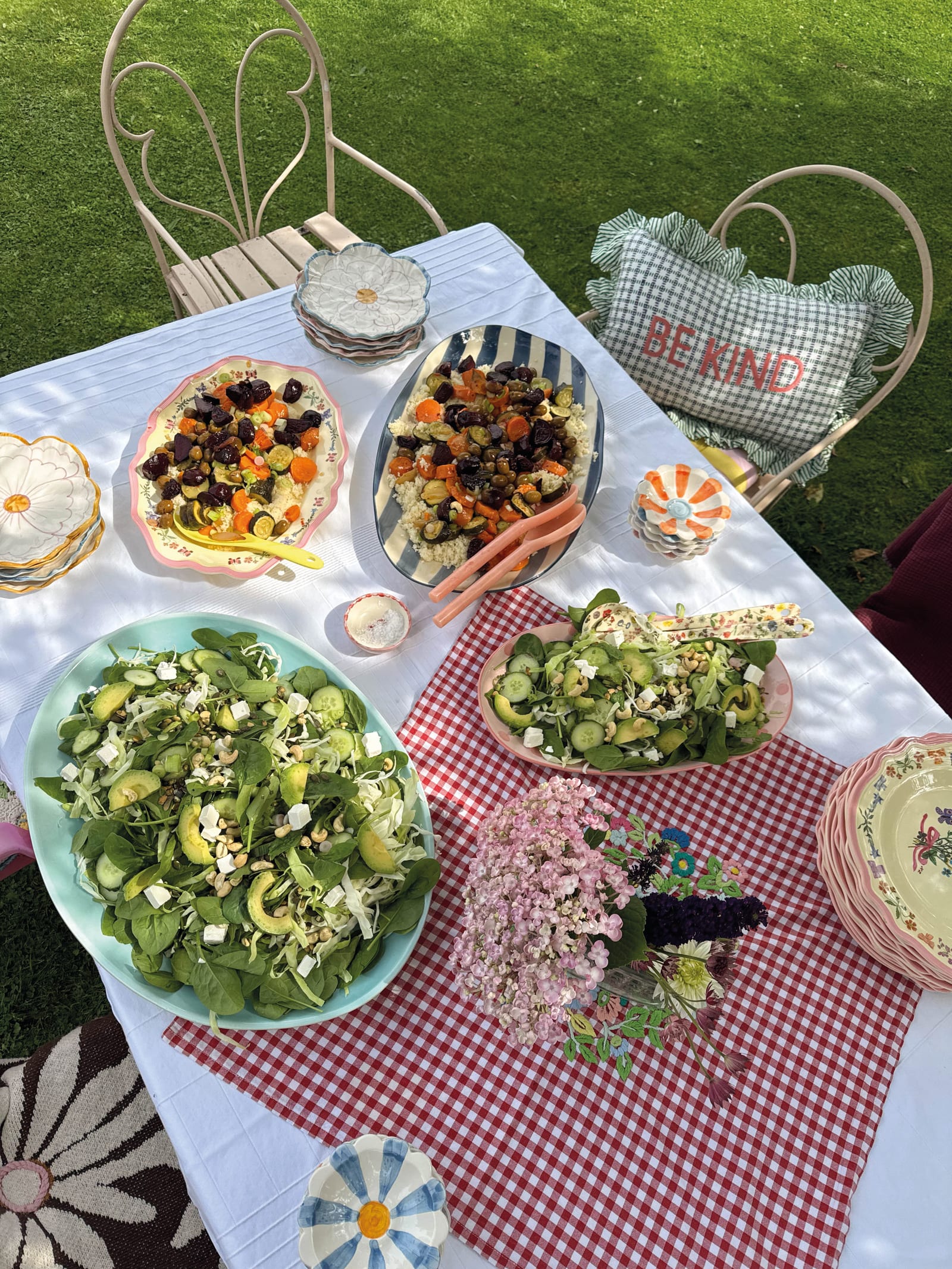 Table de jardin dressée avec nappe blanche, plats à motifs floraux, salades et couscous coloré dans le jardin