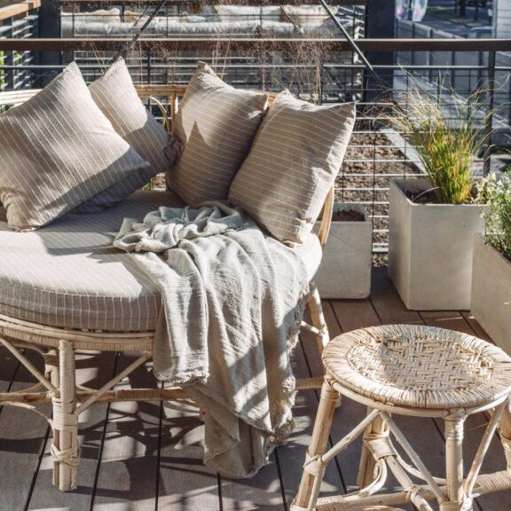 Banquette ronde en rotin sur un balcon, garnie de coussins rayés beiges et d’un plaid en lin, avec un tabouret tressé et des plantes en bacs