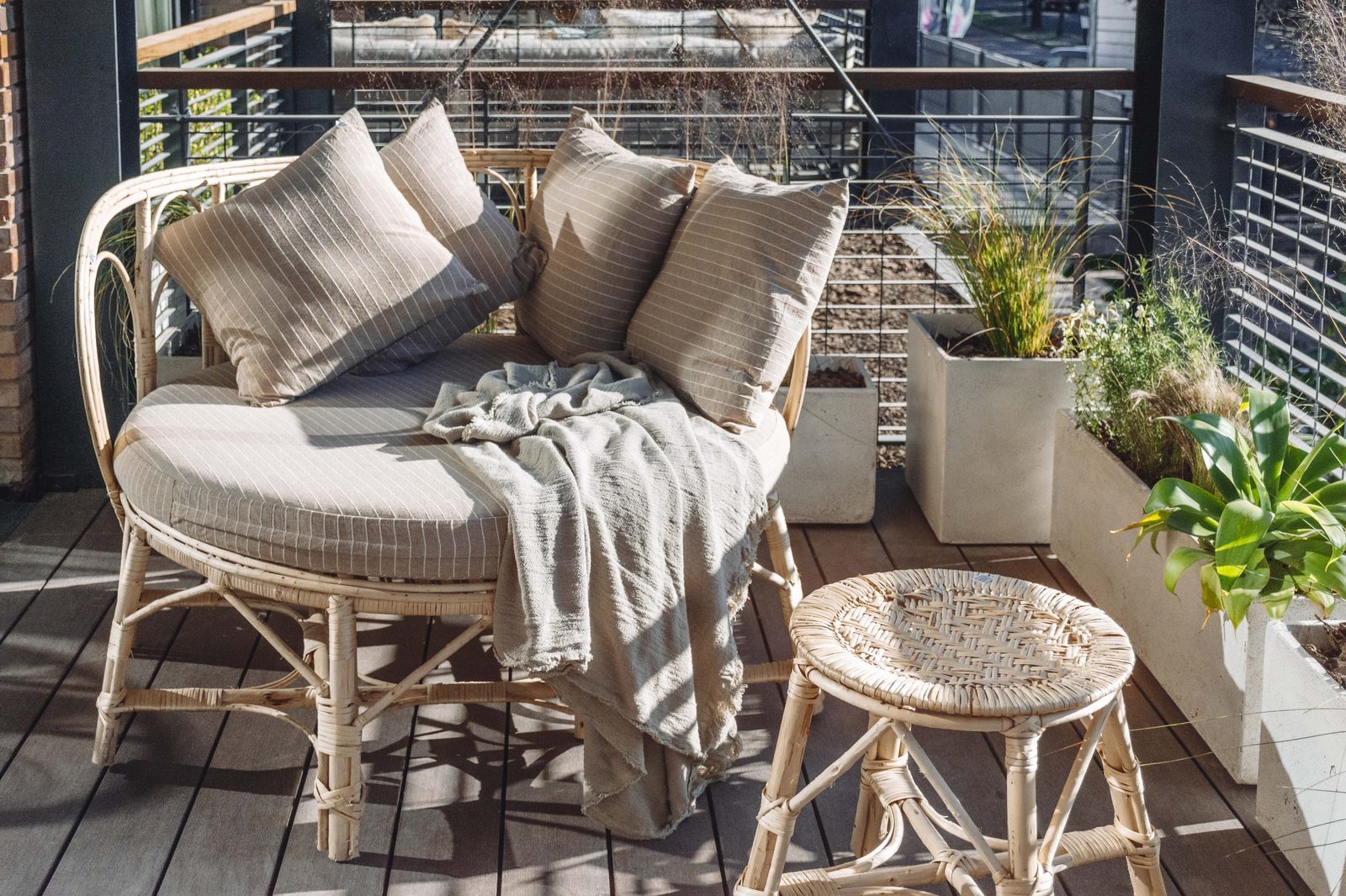Banquette ronde en rotin sur un balcon, garnie de coussins rayés beiges et d’un plaid en lin, avec un tabouret tressé et des plantes en bacs