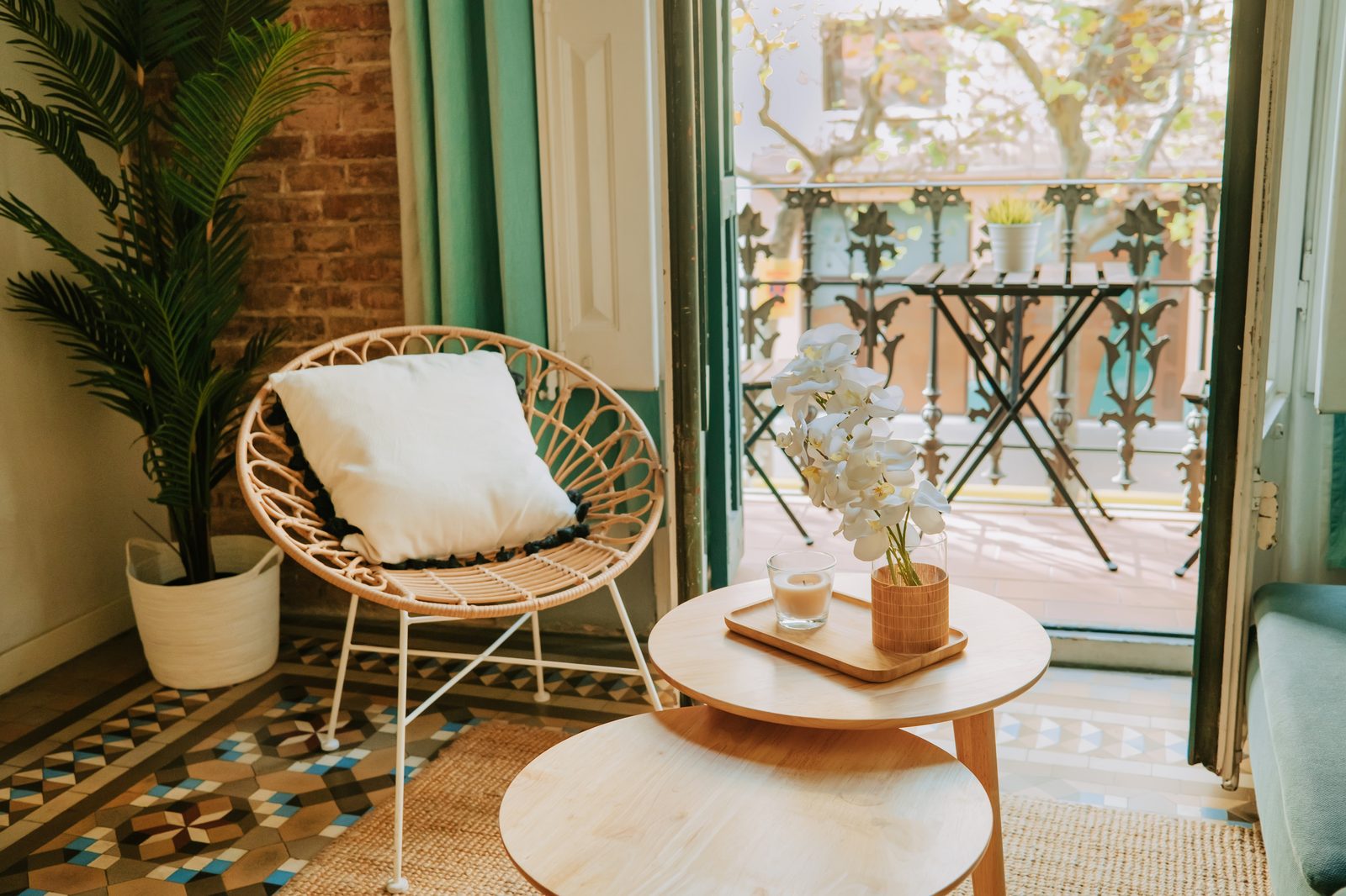 Pour aménager un petit balcon : fauteuil rond en rotin avec coussin blanc, tables gigognes en bois et sol en carreaux de ciment devant une porte-fenêtre ouverte