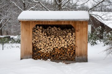 Bûcher en bois sous un toit enneigé, rempli de bûches fendues et empilées, au milieu d’un jardin couvert de neige