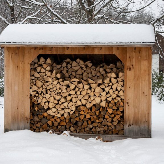 Bûcher en bois sous un toit enneigé, rempli de bûches fendues et empilées, au milieu d’un jardin couvert de neige