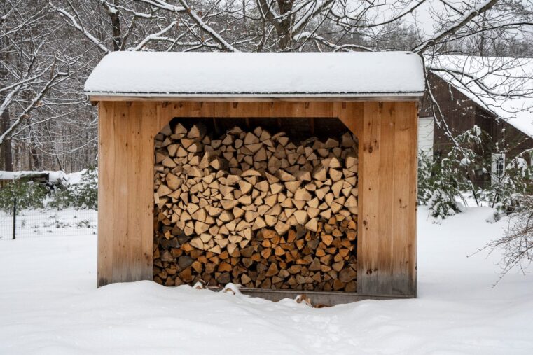 Bûcher en bois sous un toit enneigé, rempli de bûches fendues et empilées, au milieu d’un jardin couvert de neige
