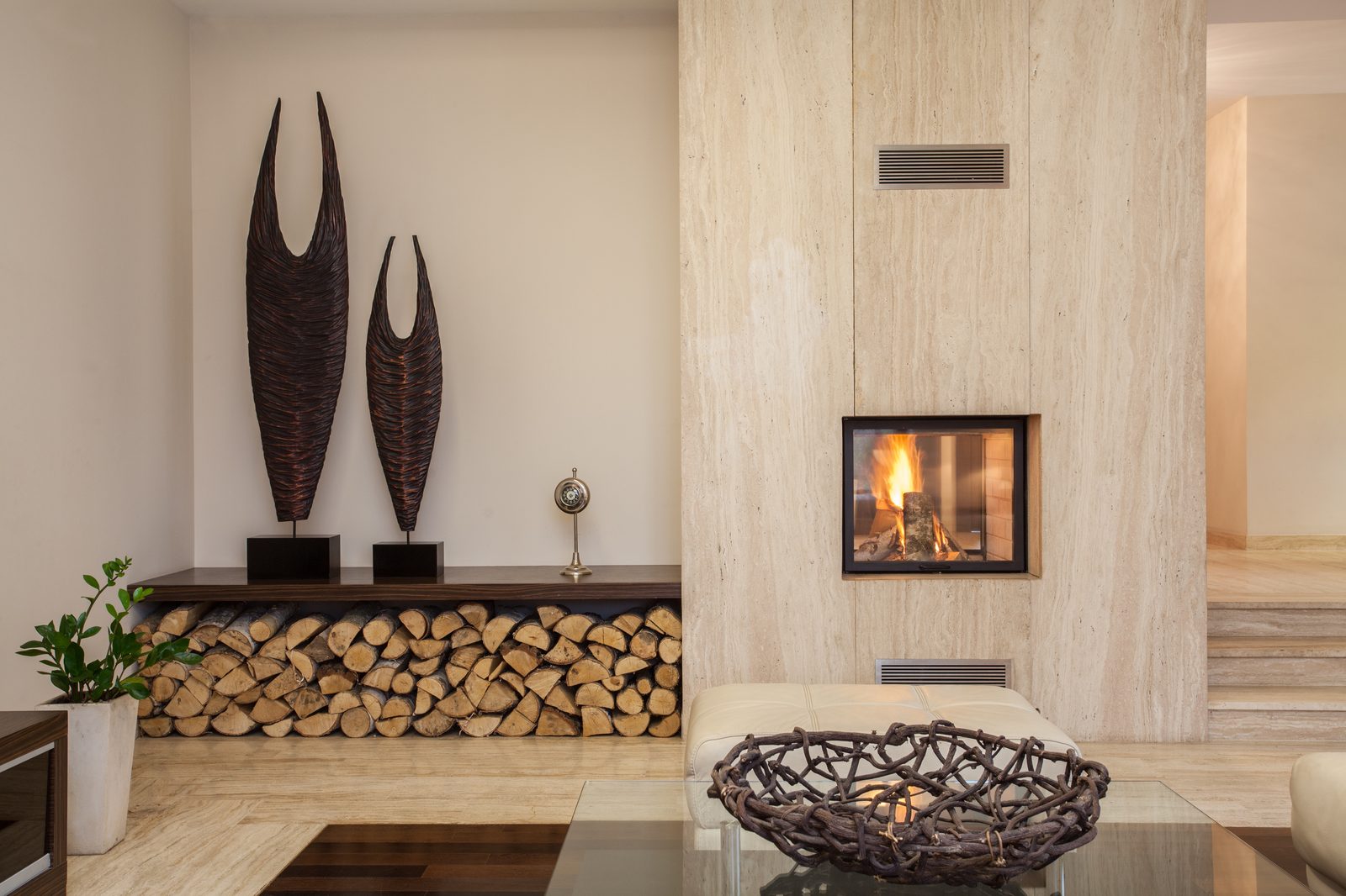 Salon clair avec cheminée encastrée dans un mur en travertin et bûches rangées dans une niche sous une tablette en bois sombre