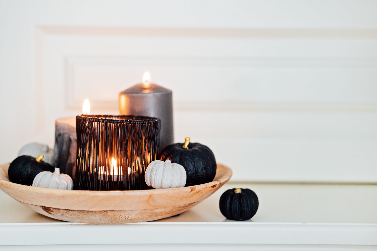 Plateau en bois avec un photophore noir, des bougies grises et de petites citrouilles noires et blanches devant un mur blanc