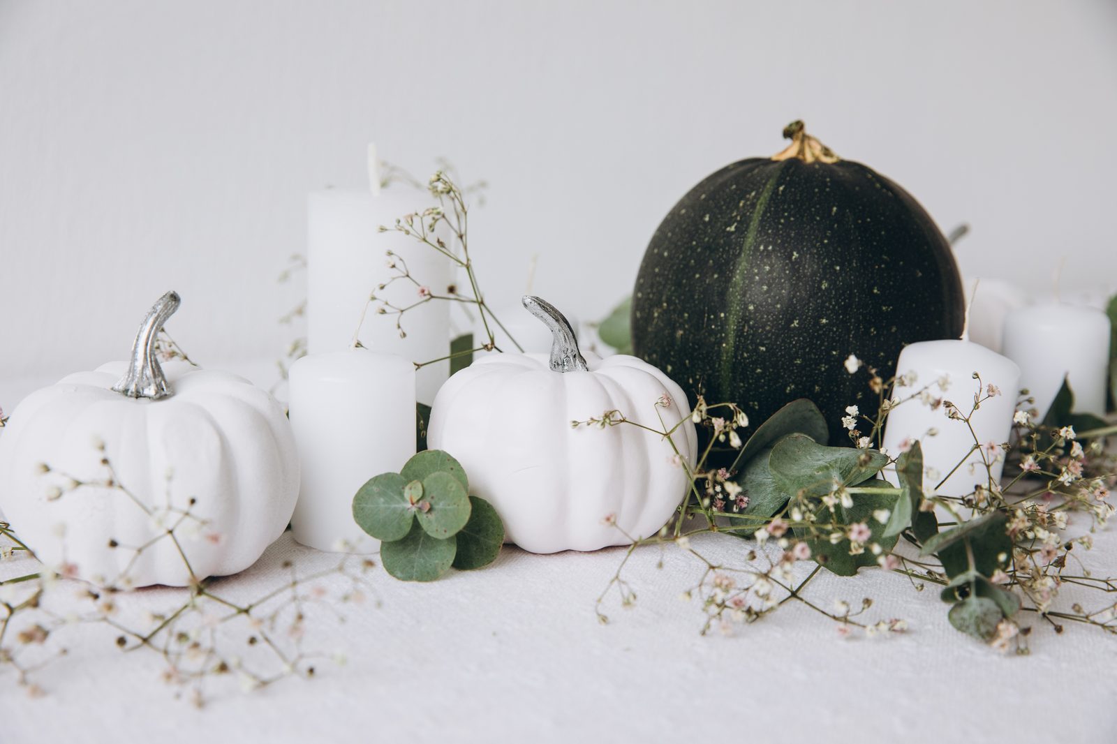 Décoration d’Halloween : citrouilles blanches à queue argentée, citrouille verte foncée, bougies blanches, eucalyptus et gypsophile sur une table claire