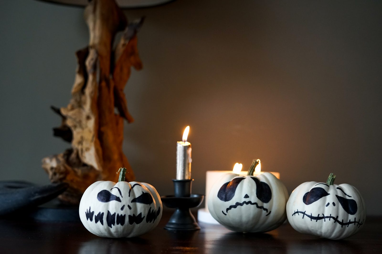 Trois petites citrouilles blanches aux visages peints en noir, alignées sur une table sombre autour d’un bougeoir noir et d’une bougie allumée