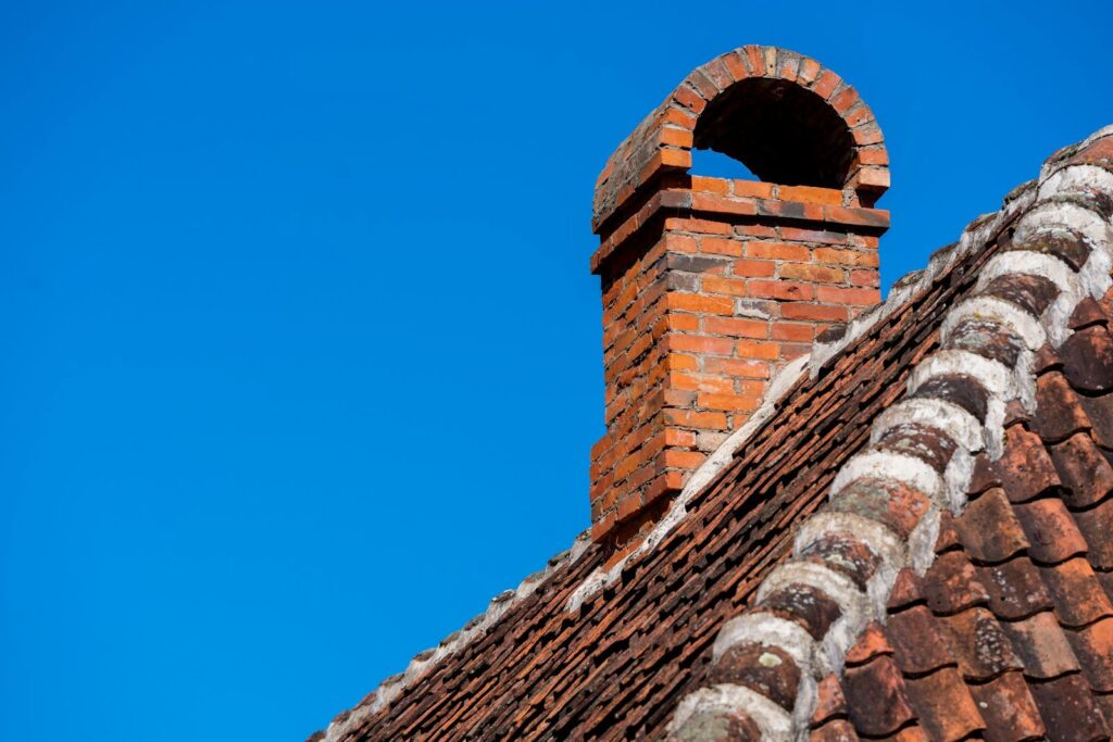 Souche de cheminée en brique coiffée d'un chapeau en arc, sur un toit de tuiles anciennes sous un ciel bleu