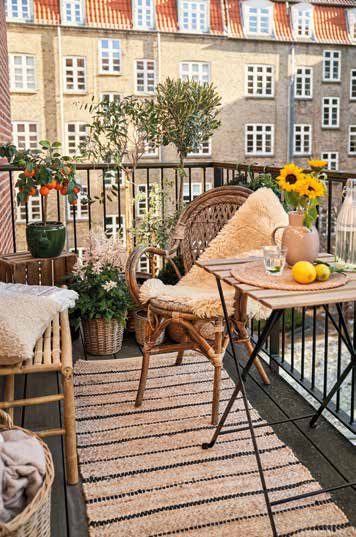 Petit balcon d'immeuble avec fauteuil en rotin couvert d'une peau, table pliante, tapis rayé et plantes en pots