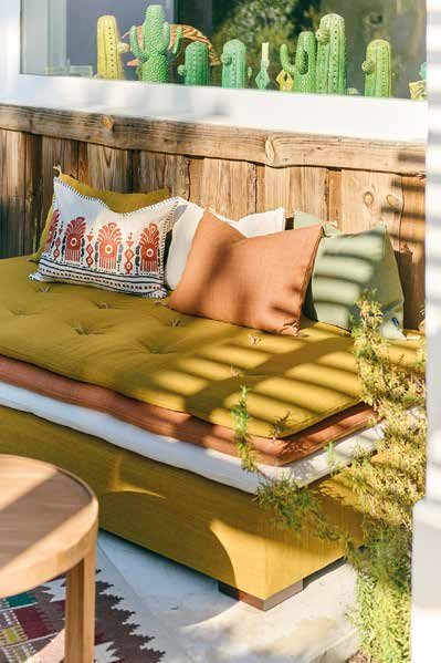 Banquette d'extérieur garnie d'un matelas moutarde et de coussins terracotta, vert et brodés, au soleil contre une palissade en bois