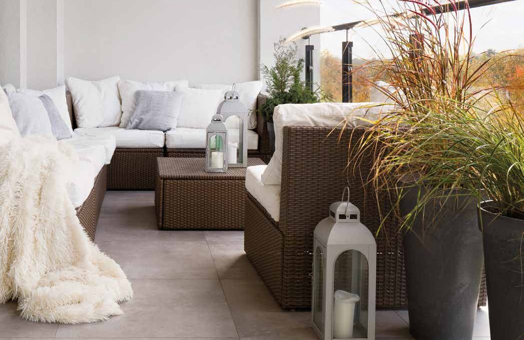 Balcon d'angle avec salon en résine tressée brune, coussins blancs, plaid à poils longs, lanternes et graminées en pots