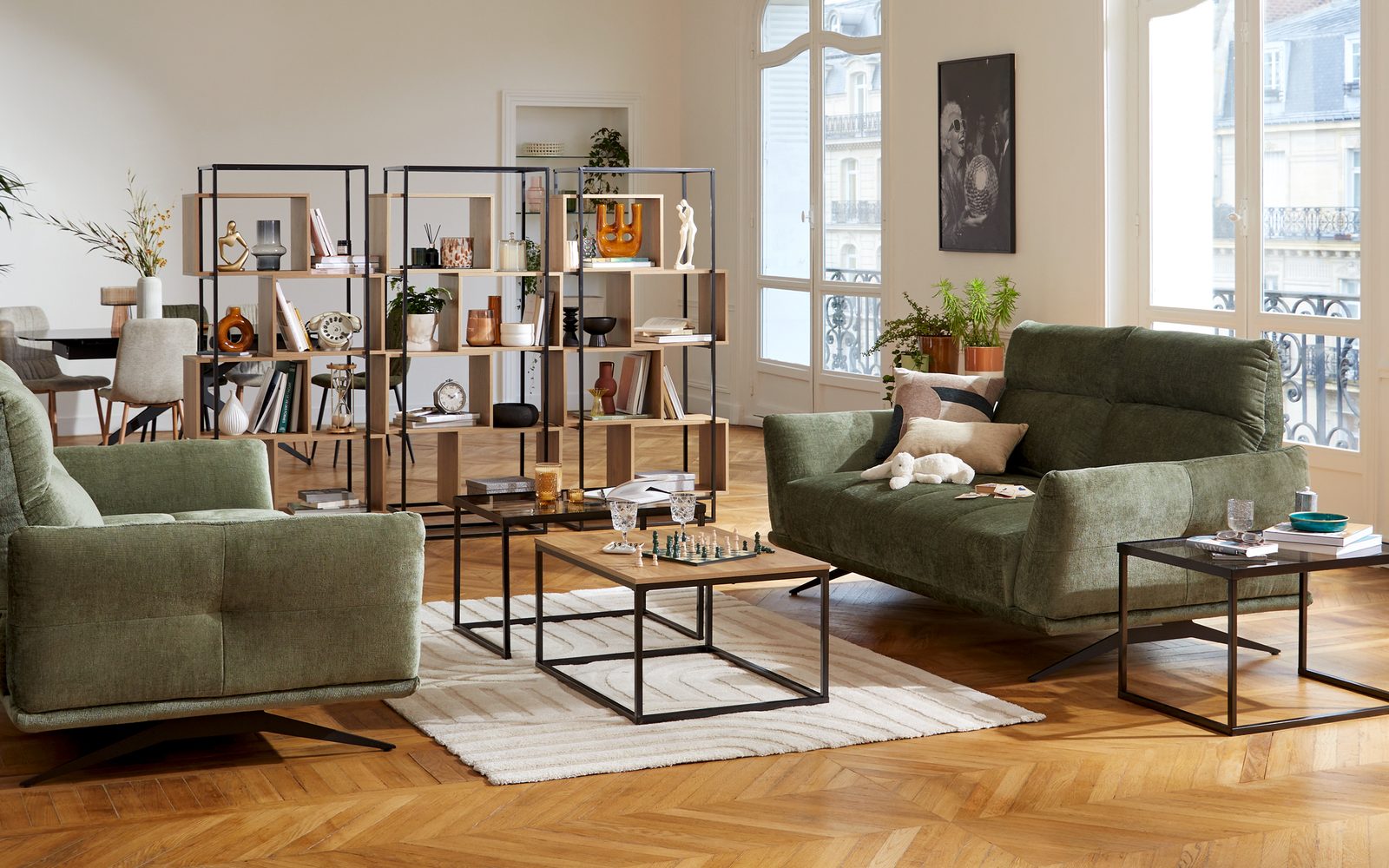 Grand séjour parisien avec canapés verts, tables basses en métal et bibliothèques ajourées en bois et métal séparant le coin repas