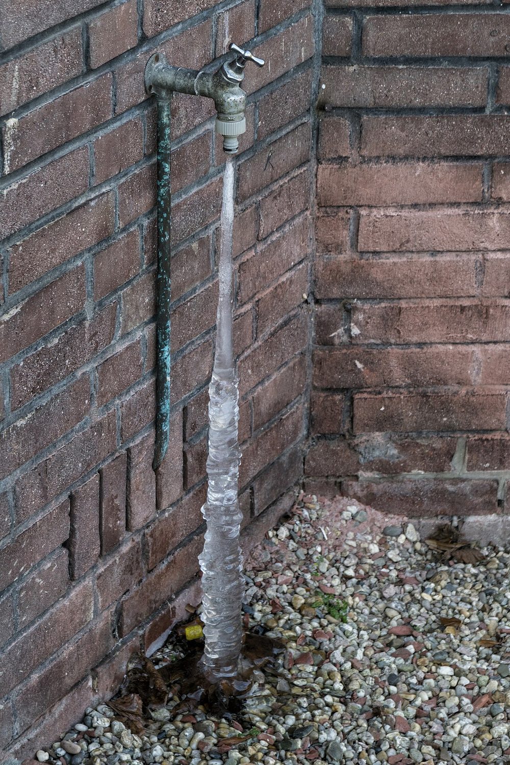 Robinet extérieur fixé sur un mur de briques, prolongé par une longue colonne de glace jusqu'au sol de gravier