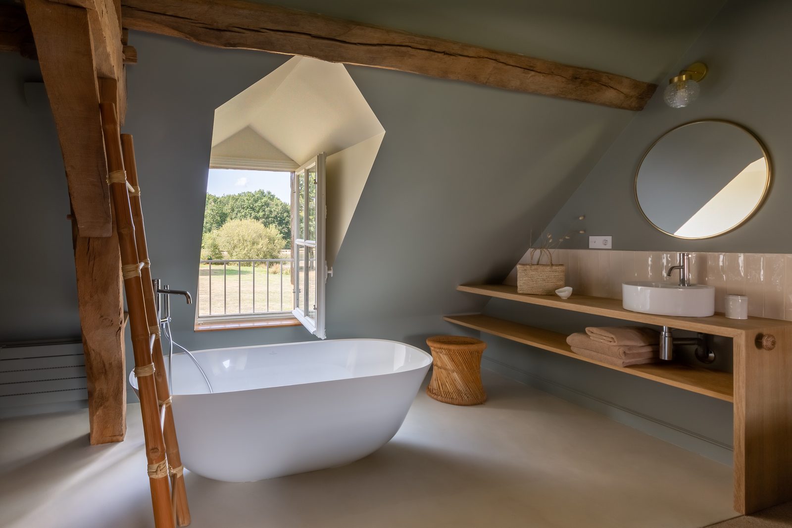 Salle de bains sous combles avec baignoire îlot, poutres apparentes et vue sur la campagne