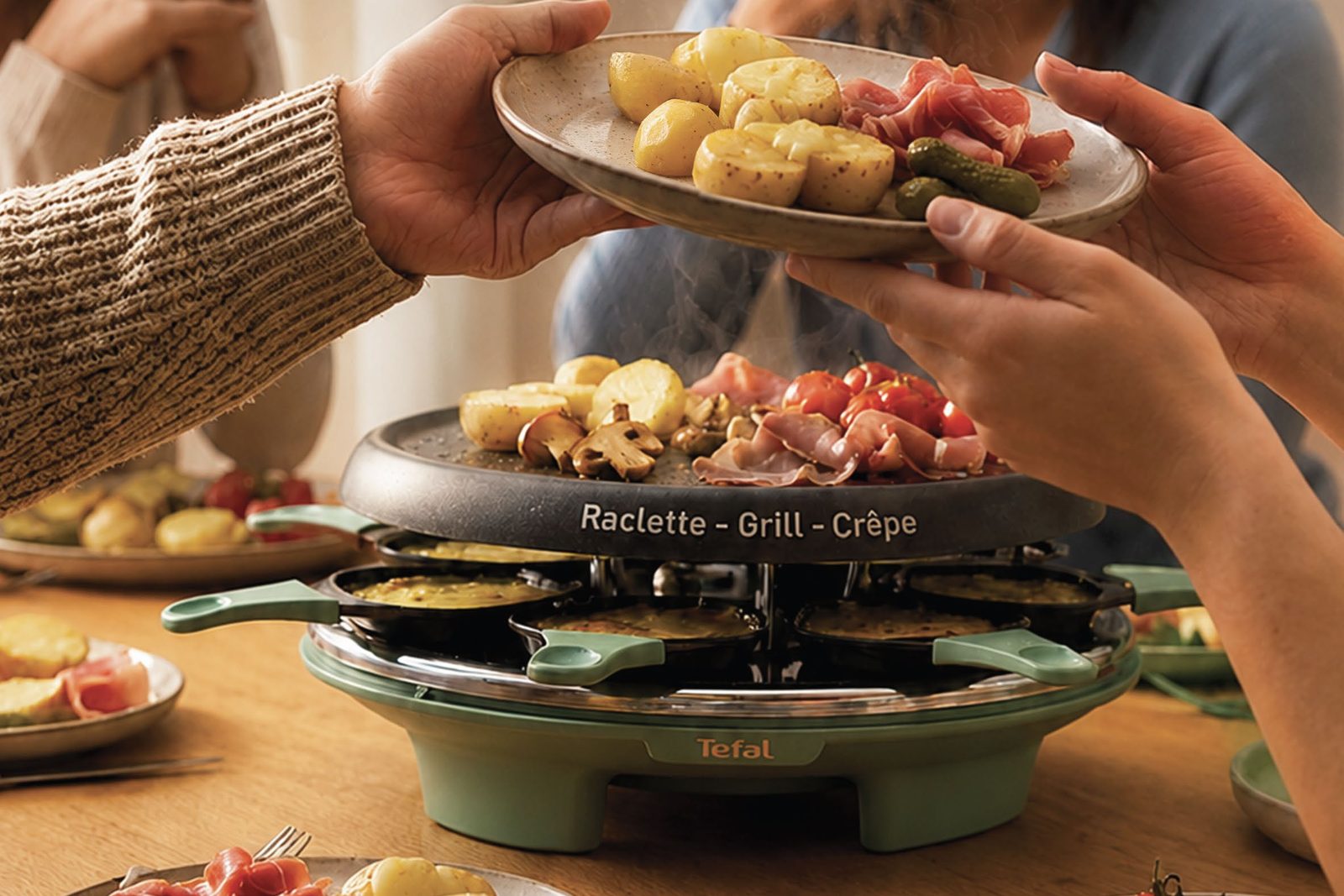 Assiette de raclette passée de main en main au-dessus de l’appareil Tefal vert sauge posé sur une table en bois