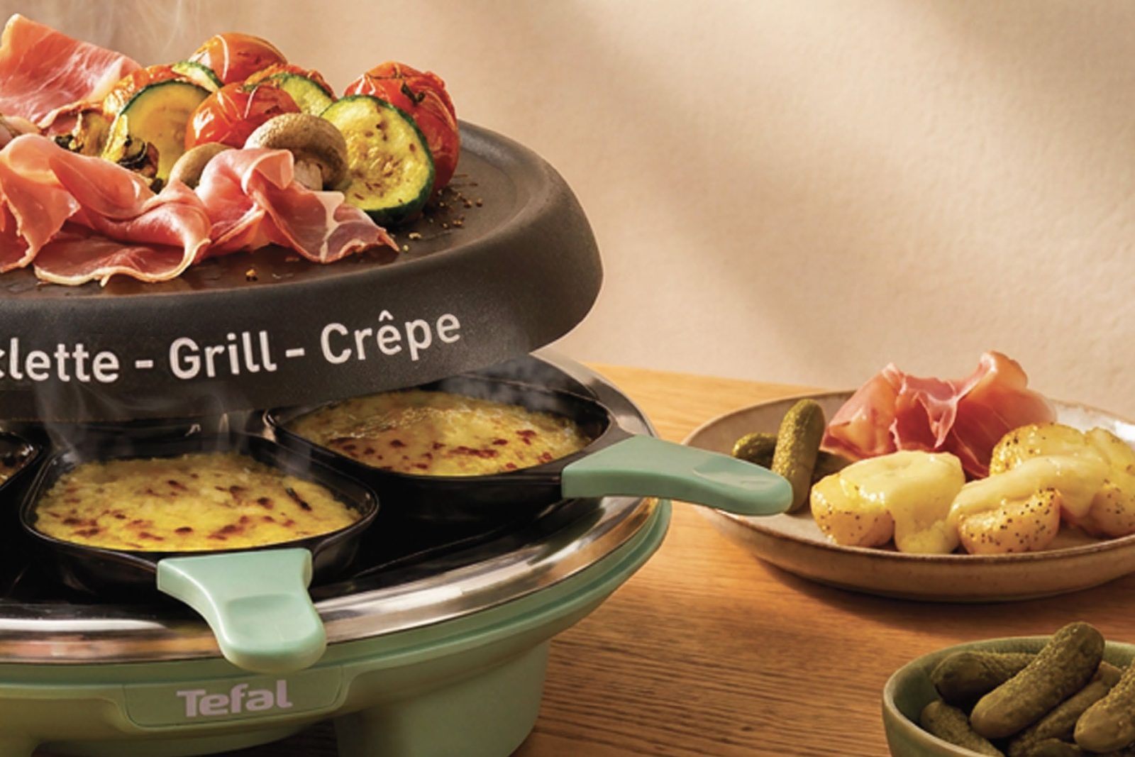 Appareil à raclette Tefal Raclette Célébration vert sauge, poêlons de fromage fondu sous une plaque garnie de jambon cru et de légumes grillés
