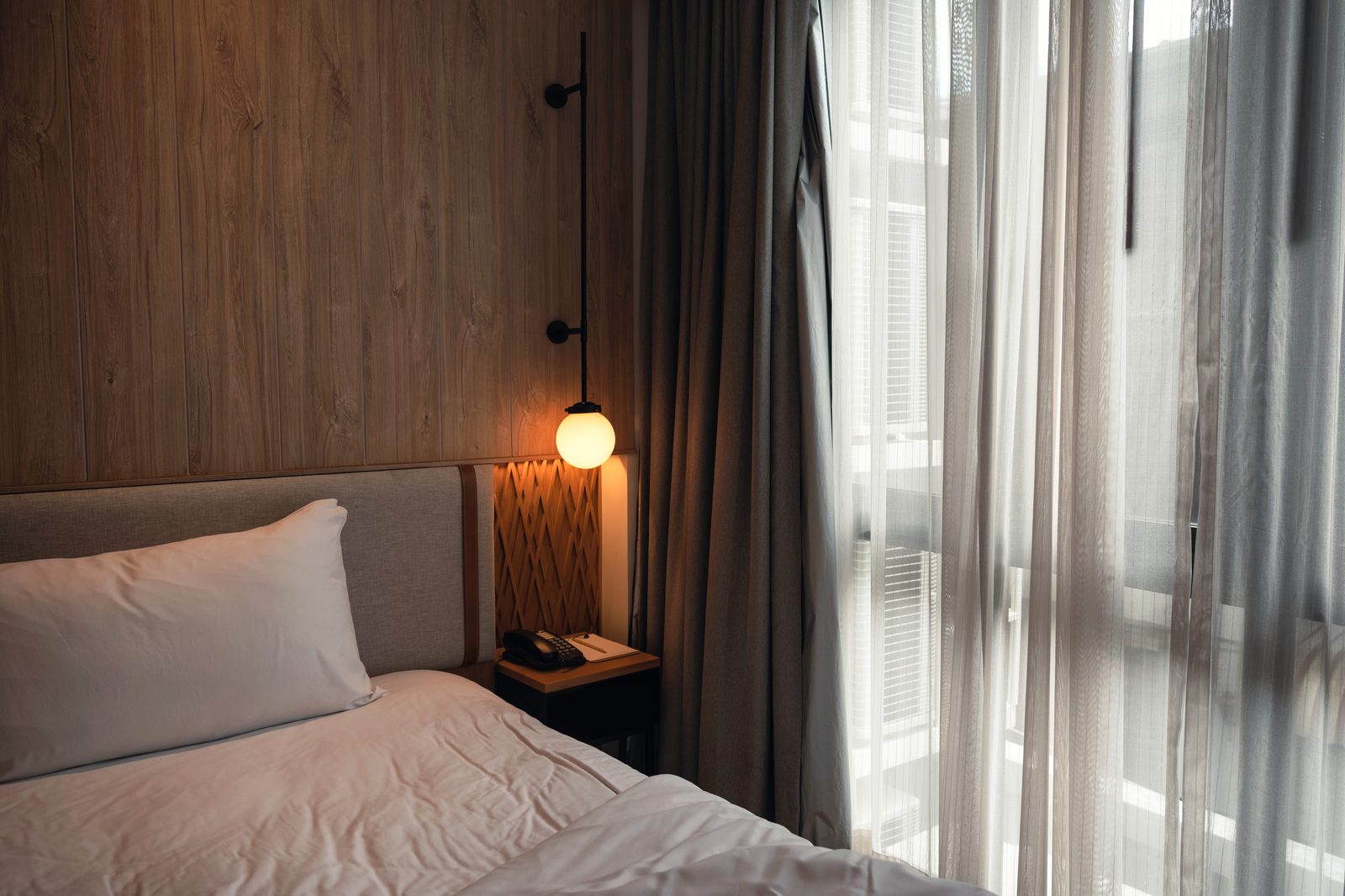 Chambre avec panneau mural en bois, applique à globe allumée, tête de lit en tissu, voilages clairs et rideaux gris