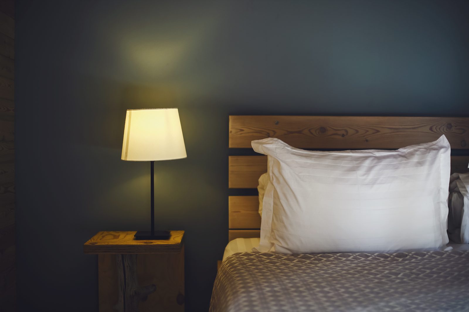 Mur bleu nuit derrière une tête de lit en bois, grand oreiller blanc et lampe de chevet à lumière chaude
