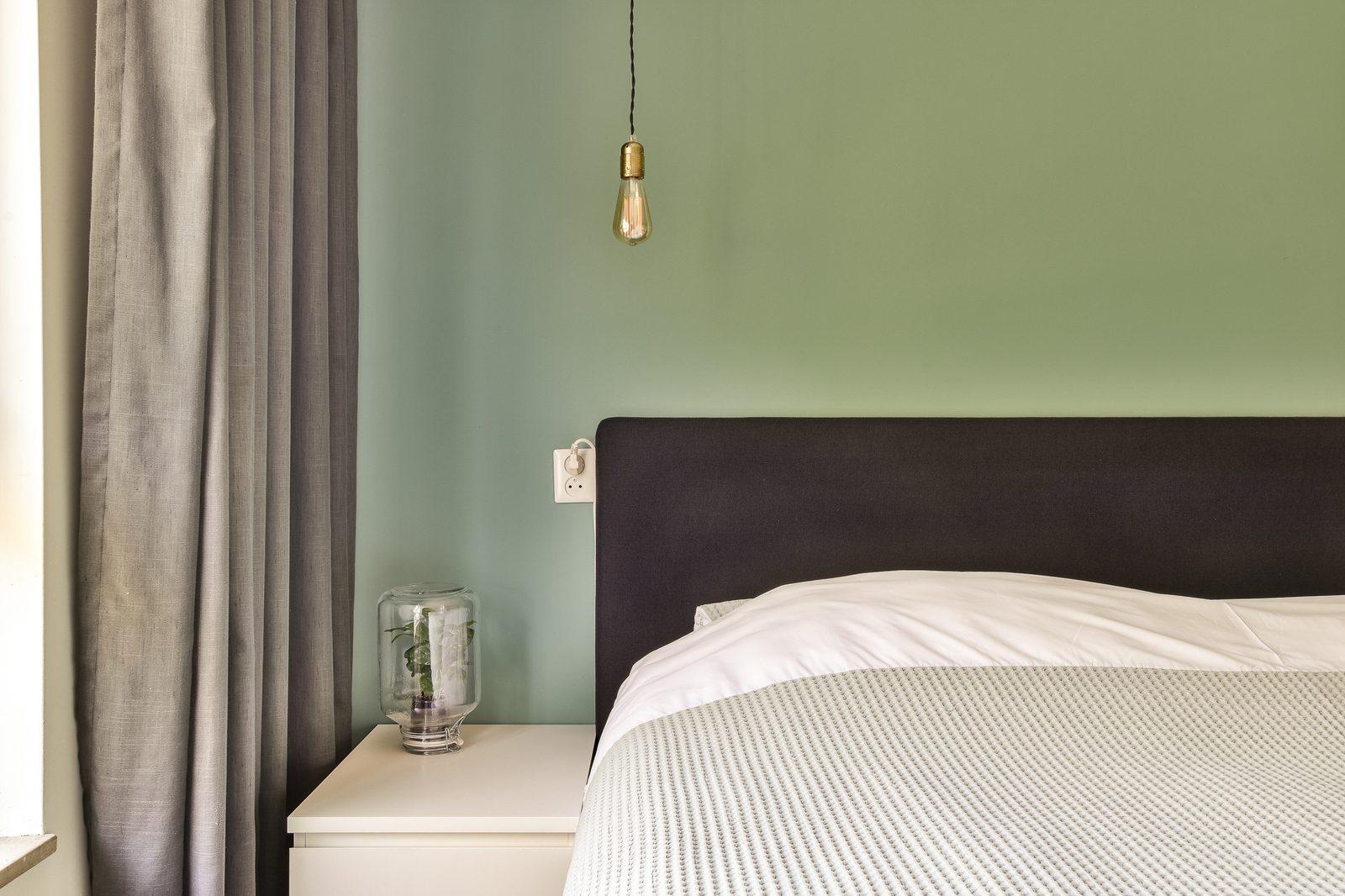 Chambre aux murs vert sauge, tête de lit sombre, lit habillé de blanc, suspension à ampoule et rideaux gris