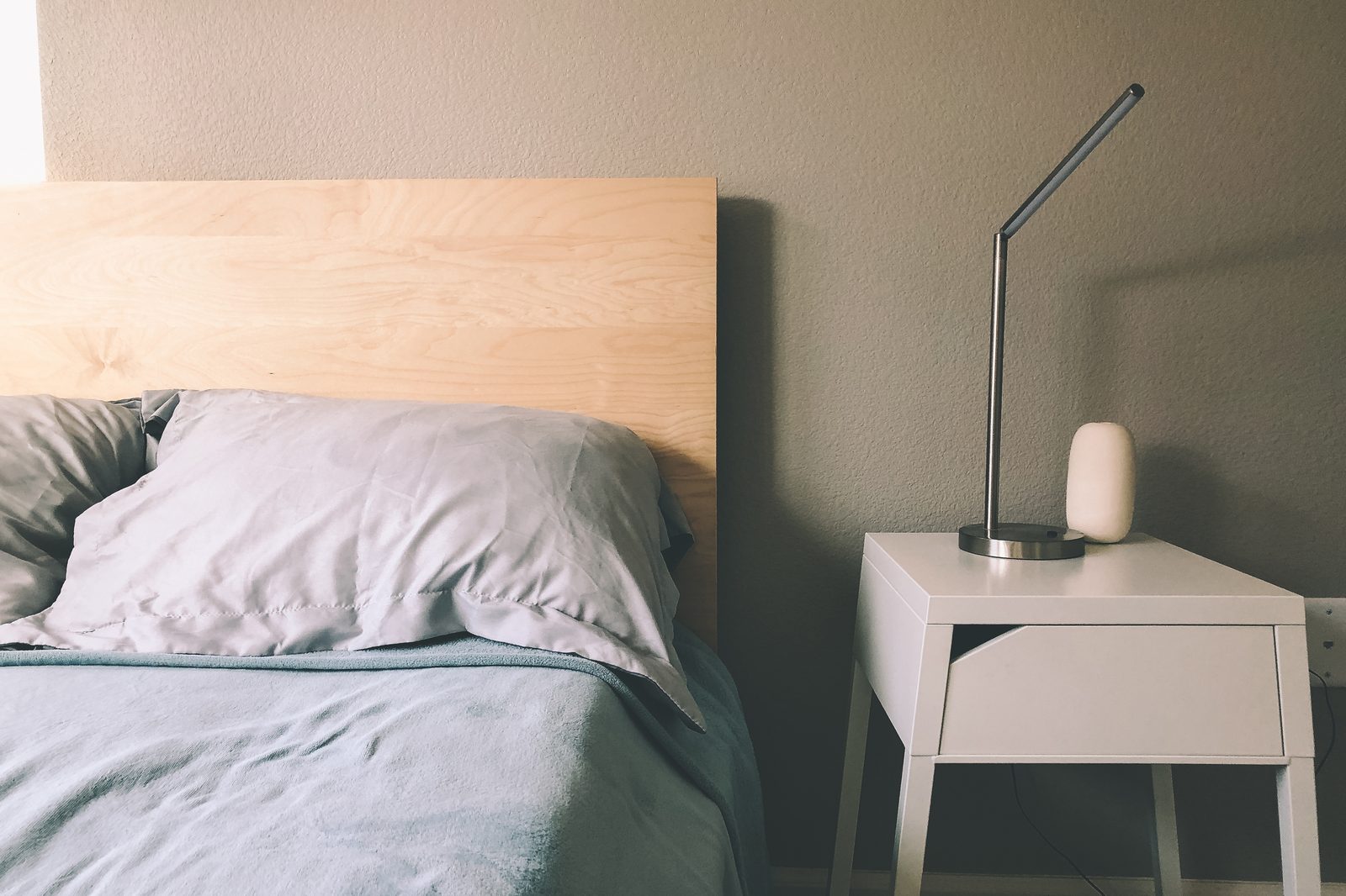 Pour aménager sa chambre pour mieux dormir : tête de lit en bois clair, linge de lit gris-bleu et table de chevet blanche avec lampe articulée