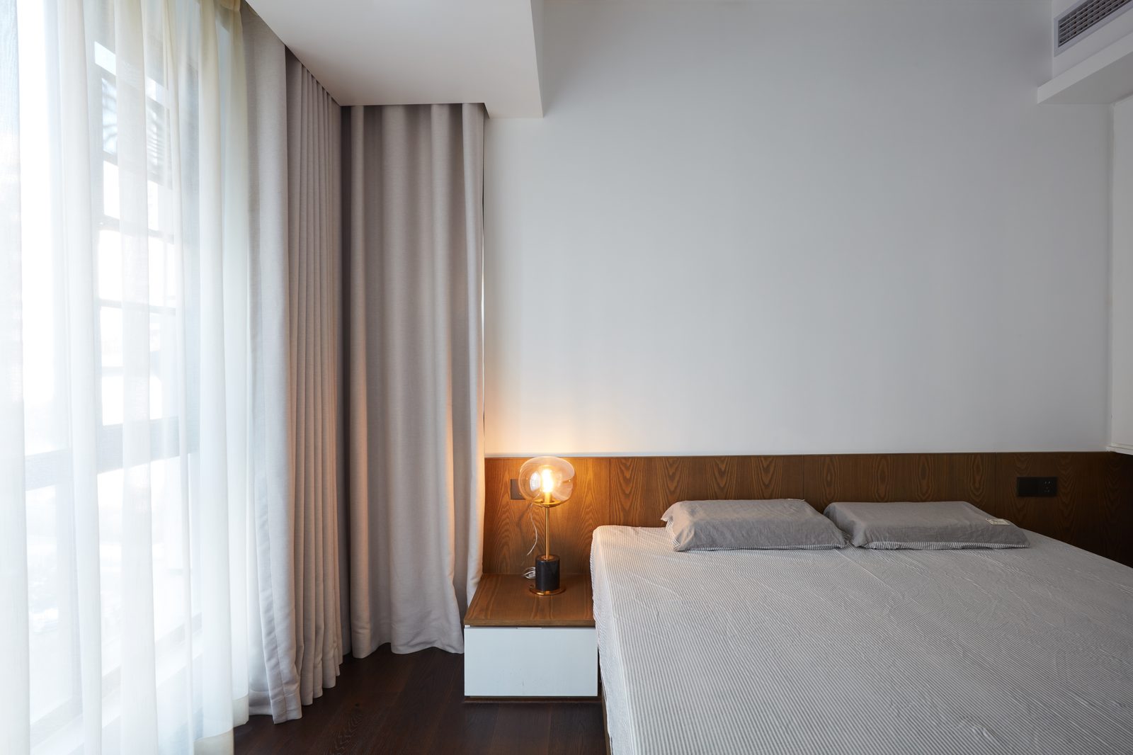Grande baie vitrée habillée de voilages blancs et de rideaux taupe, lit bas avec tête de lit en bois et lampe de chevet allumée